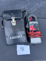 Black Hioki analog multimeter with rotary dial, clamp, attached test leads, and black carrying case labeled 'Hioki'.