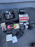 View of Sony AF-202 camera with case, Pentax lenses and lens boxes, red compact camera, Tiffen filter, film rolls, and flash unit laid out on black surface