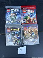 Four PS3 Lego video game cases arranged in a square on a black surface, visible titles and cover art for Star Wars III, Marvel Super Heroes, Batman 2 DC Super Heroes, and Indiana Jones 2.