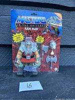 Front view of the Masters of the Universe Ram Man figure in original sealed packaging with two gray weapons included, displayed against a dark blue wall.