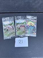Photo showing two sealed Koraidon Pokemon card packs and one holographic individual card with a colorful abstract dragon figure, placed on a wooden surface with a handwritten '21' note in front.