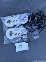 Photo shows two original SNES wired controllers, one in bag, one with taped wire, placed on dark background with lot number 34 visible.