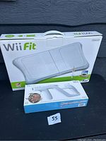 Photo showing boxed Wii Fit Balance Board and sealed Wii Zapper accessory together on a surface outdoors against dark siding.