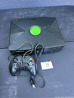 Original Xbox console and one wired controller on display, top view showing green Xbox logo on console and buttons on controller.