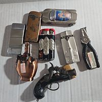 Photo of 7 vintage novelty lighters arranged on a white surface: car-shaped lighter with pin-up image, pistol lighter, hand-shaped lighter, black eagle emblem lighter, rivet plate silver lighter, brushed metal lighter, and pliers-shaped lighter.