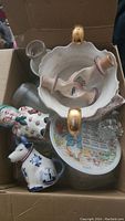 Top view of box showing assorted vintage milk bottles and various ceramic collectibles including figurines, a cow-shaped container, and decorative plates.