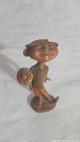Full view of the carved wooden boy figurine standing upright on a rounded base, holding a flower, with the shirt color faded.