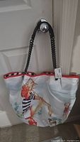 Front view of the tote bag with illustration of a woman holding a drink, black braided handles, red trim