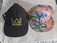 Two hats side by side, multicolor Yankees 59FIFTY fitted cap size 7 3/8 on right, black cap with gold 'KING' and crown on left.