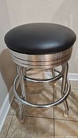 Front angled view of barstool showing padded black vinyl seat, chrome frame with ribbed detail beneath the seat, and circular footrest. Minor wear visible on chrome parts.