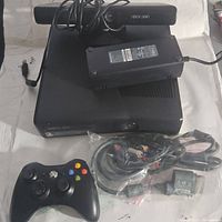 Xbox 360 console with one black wireless controller, power brick, and AV cables laid out on a white surface.