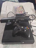 Xbox 360 console with Kinect sensor and wireless controller, showing front and top with cables attached and one game case behind.