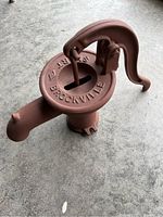 View of the hand pump top showing the lever, top round plate with 'SMART BROCKVILLE' markings, and the spout on the side.