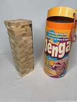 Stacked wooden Jenga blocks alongside original cylindrical yellow storage container with carrying handle and colorful printed instructions.