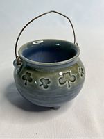 Miniature ceramic pot with shamrock cut-outs and metal handle, shown from front angle with visible glaze and three small feet.