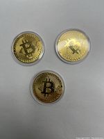 Three gold-tone Bitcoin novelty coins in clear plastic protective cases, placed on a white background.