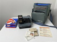 Polaroid Impulse camera, two unopened expired film packs, accessories, instruction sheet, and camera case all laid out on white surface.