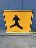 Front view of the yellow road merge sign against a blue wall showing the complete symbol and some mounting holes.