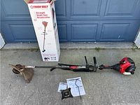 Full assembled brushcutter and accessories laid out beside original Troy-Bilt box against garage door