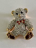Close-up photo of the vintage Swarovski teddy bear brooch showing the clear crystals covering the bear's body, the black crystal eyes, red enamel bow tie, and red enamel paw pads, along with the gold-tone finish.