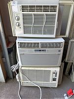 Three window air conditioners stacked in garage: top and bottom identified as GE models, middle as Electrolux model, cords visible.