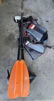 Photo of orange Intex branded dual-blade oars alongside trolling motor and life vests on concrete floor.