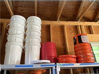 Thirteen white plastic buckets stacked in two columns with orange and red lids stacked beside them on a metal rack.
