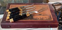 Photo showing six carving knives and one carving fork with black handles and gold plated accents spread on a wooden box