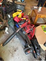 View of assorted leaf blowers, chainsaws, and multiple empty gas cans piled on a garage floor.