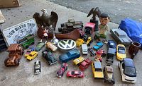 Overall collection showing various vintage toy cars, bobblehead figures, Avon bottles, an eagle figurine, and miniature collectible items on outdoor surface with visible dirt.