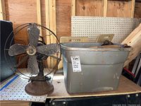 Wide shot showing the Graybar fan next to a metal bin filled with various wood planes on a workbench in a garage setting
