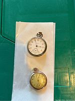 Two pocket watches placed on a white sheet on a green surface, showing both watches in full.