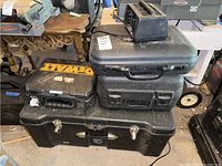 Photo shows stacked black tool trunk and several black plastic tool cases including one DeWalt branded case, along with a Panasonic VHS camcorder battery charger on top.
