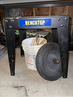 Black metal benchtop stand with white plastic bucket inside containing wooden mallets. Tire wheel leaning against the stand.