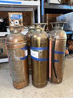 Four vintage metal fire extinguishers grouped together on concrete floor, showing tops and body details with blue tape date label.