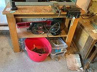 Wood work table with shelves holding power tools and hardware, with metal vises mounted on the top edge.
