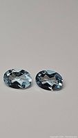Pair of natural oval cut sky blue topaz gemstones displayed on plain background showing facets and clarity.