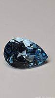 Pear cut deep Swiss blue faceted gemstone showing surface details and facets under direct light.