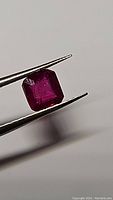 Close-up image of natural ruby gemstone held in tweezers, showing square cut and rich blood red color