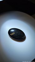 Close-up photo of a polished black obsidian oval cabochon gemstone with golden sheen effect under direct light.