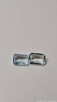 Pair of rectangular emerald cut sky blue topaz gemstones on a plain neutral background, showing clarity and color.
