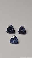 Three natural trillion cut tanzanite gemstones displayed on neutral background showing their purple-blue coloration and faceted cuts.