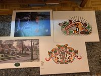 Four art prints laid out on floor showing various Indigenous artworks, architectural street scene, and modern atmospheric artwork.