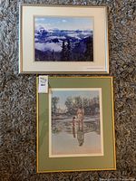 Top view showing both framed pieces on carpeted floor, with visible tags, one artwork shows a mountain landscape and the other a man standing in water with a child.
