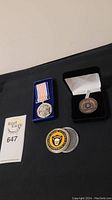 Four items on black surface including two medals, one with blue and white-striped ribbon, and three coins, one with yellow and black tiger emblem.