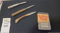 Three folding knives and a Seber Tool multi-tool kit in a plastic case on black surface.