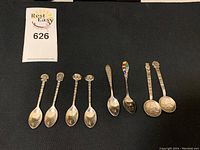Eight assorted sterling silver collector spoons displayed on black fabric showing varied decorative handles including patterned heads, enamel, and embossed scenes.