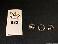 Three sterling silver rings displayed on black fabric: bold abstract wave ring, plain band, small rounded thumb ring.