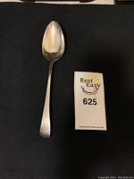 Top view of sterling silver spoon placed on a dark surface next to a lot number card.