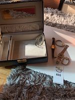 Two wrist watches and jewelry box shown with one watch having a brown woven strap and gold dial, another watch with metal band, and open jewelry box.